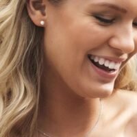 orhangen_halsband_blomdahl__7330981540862_7330981590461_15-1233-59_33-2433-0659.jpg صورة مقربة لامرأة مبتسمة ترتدي أقراطًا من اللؤلؤ وقلادة رقيقة، مما يُظهر تعبيرها المبهج وإكسسواراتها الأنيقة.