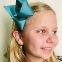 orhangen_titan_blomdahl_7330981158166_15-1255-021.jpg A young girl smiling with a turquoise hair bow and yellow earrings, showcasing a cheerful expression and light blonde hair.