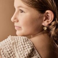 orhangen_titan_blomdahl_7330981158166_15-1255-022.jpg A young girl wearing heart-shaped earrings, showcasing a close-up view of the jewelry and her profile with natural lighting.