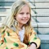 orhangen_titan_blomdahl_7330981158166_15-1255-02_b.jpg A young girl with blonde hair wearing a pineapple-patterned jacket, sitting on stone steps, smiling brightly with blue eyes.