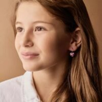 orhangen_titan_blomdahl_7330981158173_15-1255-03.jpg A young girl wearing heart-shaped pink earrings, showcasing a stylish accessory for children. Perfect for adding charm to any outfit.