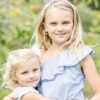 orhangen_titan_blomdahl_7330981158180_7330981521380_15-1255-05_15-1249-06.jpg Two young girls wearing matching striped outfits, one with blonde hair and blue eyes, the other with curly blonde hair, smiling outdoors.