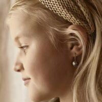 orhangen_titan_blomdahl_7330981158531_15-1255-12.jpg A young girl with blonde hair wearing heart-shaped earrings and a braided headband, showcasing a side profile view.