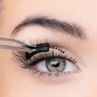 9045-e1655732839928 Close-up image of a person applying false eyelashes using tweezers, showcasing long, voluminous lashes and a detailed eye.