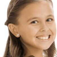 orhangen_nickelfri_plast_blomdahl_7330981510643_15-01121-01_a.jpg A young girl smiling while wearing heart-shaped earrings, showcasing a joyful expression and light brown hair.