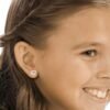 orhangen_nickelfri_plast_blomdahl_7330981510643_15-01121-01_b.jpg Heart-shaped diamond earrings worn by a smiling girl, showcasing a delicate and charming design suitable for children.
