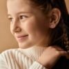 orhangen_titan_blomdahl_7330981156445_15-1424-24.jpg Close-up of a young girl smiling, showcasing delicate stud earrings and a braided hairstyle, with a soft background.